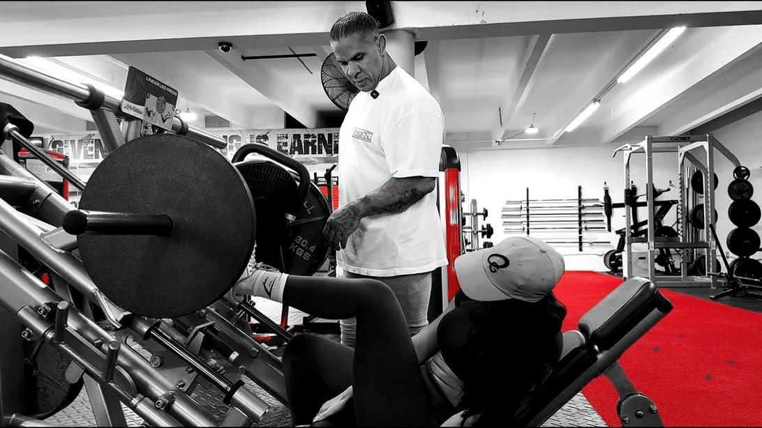 Omar corrige la técnica de una clienta en la prensa de piernas, sobre el suelo rojo del gimnasio